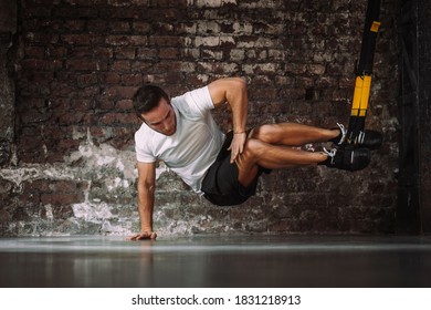 Powerful Sportsman Balancing On Arm While Doing Exercise With TRX Ropes Against Rough Brick Wall In Gym