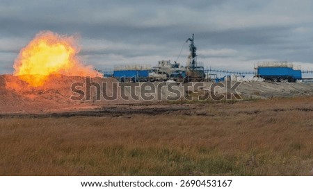 A powerful torch when burning gas coming out of a well is in the foreground. In the background, blurred in the hot air, is an image of an installation for lowering a flexible pipe into a wel