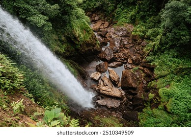 A powerful waterfall cascades down into a rocky gorge surrounded by dense, lush greenery. The water’s force creates a misty spray as it crashes onto the large boulders below, adding to the dramatic. - Powered by Shutterstock