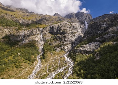 A powerful waterfall descends from a steep, rocky cliff, surrounded by patches of green vegetation and rugged rock formations. - Powered by Shutterstock