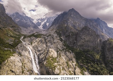 A powerful waterfall descends from a steep, rocky cliff, surrounded by patches of green vegetation and rugged rock formations. - Powered by Shutterstock