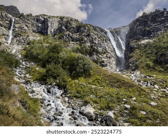 A powerful waterfall descends from a steep, rocky cliff, surrounded by patches of green vegetation and rugged rock formations. - Powered by Shutterstock