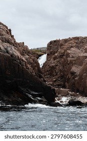 A powerful waterfall drops between steep rocky cliffs into a fast moving river, creating a dramatic natural landscape under an overcast sky with distant