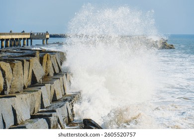 Powerful wave crashes against the rocky shore near Cabrillo Beach Pier, creating an impressive splash, with the pier and distant beachgoers visible in the background.