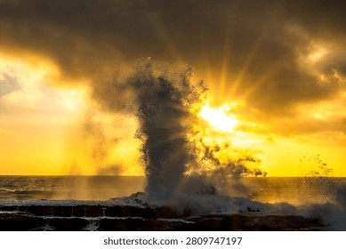 A powerful wave explodes into a spray of water against dark rocks as the sun sets, casting golden light through dramatic clouds