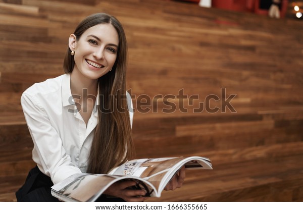 Powerful woman\
and business concept. Alluring young caucasian girl sitting on\
stairs, having lunch break, reading fashion or design magazine,\
smiling camera, relax in co-working\
space