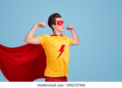Powerful young man in superhero costume with waving red cape showing biceps while being ready to save world against blue background