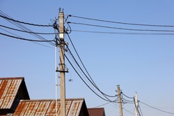 Powerline Posts With Electrical Wires And Capacitors Above Old Roofs On Blue Sky Background. Electricity Transmission Line, Power Supply In Village