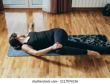 Practicing Reclined Spinal Twist Pose: Beautiful woman in sportswear doing spine rotation exercise on a gray exercise mat in the living room at home