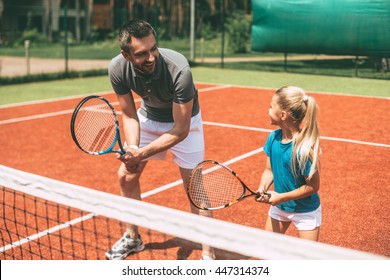 Practicing tennis. Cheerful father in sports clothing teaching his daughter to play tennis while both standing on tennis court 