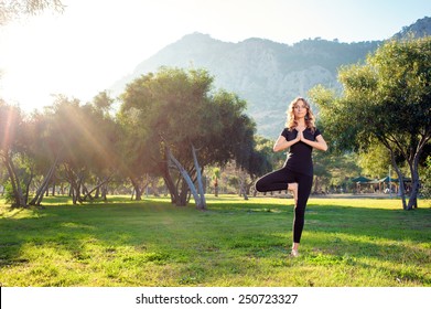 Practicing yoga in the morning, with trees, mountains and sun ray in the background. Attractive young caucasian woman standing in yoga pose on the grass.