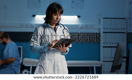 Practitioner doctor holding tablet computer checking patient report while typing medical expertise during night shift in hospital office. Physician woman in white coat working at health care treatment