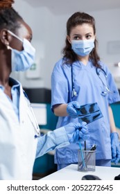 Practitioner Nurse Giving Xray To African American Radiologist Doctor Examining Lungs Disease During Clinical Appointment In Hospital Office. Medical Team With Protective Face Masl Against Covid19