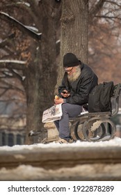 Prague, Czech Republic. 02-23-2021. Old Homeless Man Is Eating And Reading The Newspaper Sitting On A Bench In The City Center Of Prague On A Cold Winter Day.