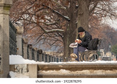 Prague, Czech Republic. 02-23-2021. Old Homeless Man Is Eating And Reading The Newspaper Sitting On A Bench In The City Center Of Prague On A Cold Winter Day.