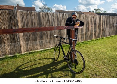 Prague, Czech Republic - Cca May, 2020: Smiling Bearded Man With Facemask On His Ear Has Pilsner Urquell Beer In Garden Pub During Coronavirus Pandemic. Social Distancing Hipster With Cannondale Bike.