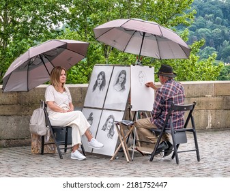 Prague, Czech Republic - June 2022: Street Artist On Charles Bridge Making A Sketch Or A Portrait To A Woman Sitting On A Chair