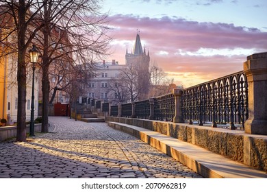 Prague, Czech Republic. Sunrise sunshine sunbeams and shadows on paving stones from promenade from river Vltava at old Winter street with trees and lantern. Tower Charles Bridge. Scenic sky clouds.
