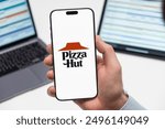 PRAGUE, CZECHIA - JULY, 2024: Pizza Hut logo on the screen of smartphone in mans hand and laptop on the background 