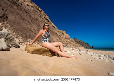 Praia da Luz in Algarve in Portugal. Beautiful long hair woman in colorful swimsuit sitting on stone and exposing herself to sun against background of rocky cliff on sandy beach on sunny day. - Powered by Shutterstock