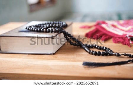 A prayer bead string rests on a closed Quran atop a wooden table; the focus is on the religious object in the foreground against a blurred background—a concept suitable for Ramadan, Hajj, and Islamic themes, featuring empty space for text.
