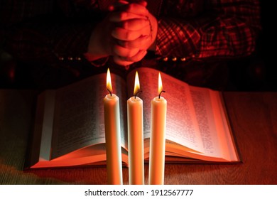 Praying hands next to bible. Man praying while reading the Bible. Crossed fingers next to book as a symbol of faith in God. Church candles in front of a praying person. Prayer hands close up.