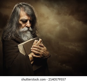Praying Monk with Bible. Prophet holding Book. Old Man Portrait with Long Gray Beard in Black Cloak over Dark Mysterious Background