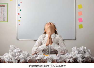 Praying School Girl With Many Wrapped Papers Around Sitting In The Classroom, Writing Composition On The Topic, Have No Idea What To Write About, Prays, Think Hard