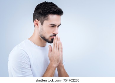 Praying. Unhappy Calm Young Man Finding Inner Peace And Harmony While Standing With His Hands Closed And Praying