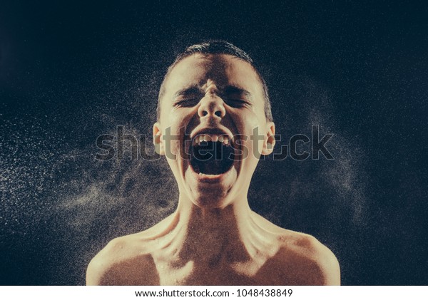 Pre teen boy without t-shirt angrily screams into a spray of water against a black background with copy space. Emotional portrait of teenager man. Toned image. screams from the front view