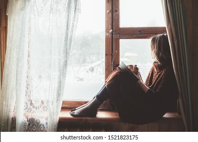 Pre teen child in warm woolen sweater seating on window sill and reading a book. Winter weekends in old log house. Cold snowy weather. Cozy homely concept.