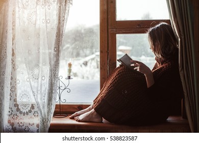 Pre Teen Child In Warm Woolen Sweater Seating On Window Sill And Reading A Book. Winter Weekends In Old Log House. Cold Snowy Weather. Cozy Homely Concept.