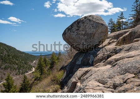 Precariously Balanced Bubble Boulder on a Mountainside Overlooking the Ocean A large, round boulder rests precariously on a smaller rock formation, offering a breathtaking view of a winding road lead