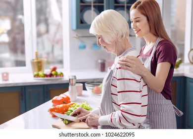 Precious bond. Charming young woman bonding to her elderly mother while cooking dinner with her together, the older woman cutting vegetables - Powered by Shutterstock