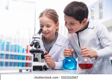 Precise look. Jolly energetic curious girl using microscope and smiling while boy holding bulbs - Powered by Shutterstock