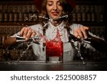 With precision, a bartender mixes a bright red cocktail, creating dynamic splashes against the backdrop of a lively bar, showcasing her deft skills and creativity.