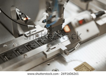 Precision robotic arm positioning electronic components onto black microchip carriers on an automated production line, with visible wiring and metallic guides