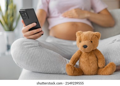 Pregnancy App For Maternity Prenatal Online Class. Pregnant Woman Using Mobile Phone With Focus On Baby Toy Teddy Bear. Nursery Shopping, Newborn Preparation, Healthcare