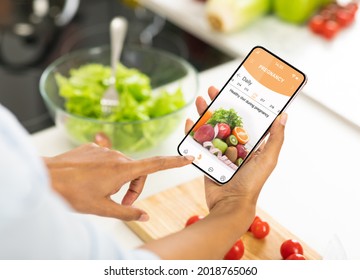 Pregnancy App. Over the shoulder of African American lady using mobile application cooking in kitchen. Female browsing nutrition guidance and recipes for healthy diet during pregnancy, prenatal care - Powered by Shutterstock