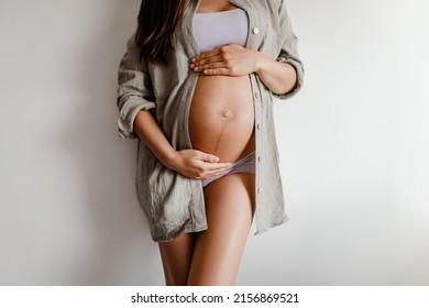 Pregnancy belly closeup. Pregnant woman wearing underwear and casual cotton shirt at home relaxing holding expecting tummy for skincare, health, lifestyle - Shutterstock ID 2156869521