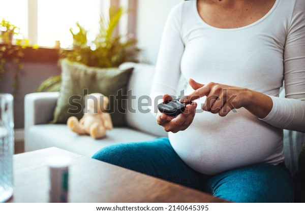 Pregnancy, health and glycemia concept - pregnant\
beautiful woman checking blood sugar level with glucometer and\
lancing device at\
home