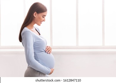 Pregnancy. Joyful Expectant Woman Touching Belly And Smiling Standing Near Window At Home. Free Space
