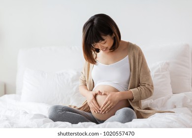 pregnancy, love, people and expectation concept - happy pregnant asian woman sitting in bed and making hand heart gesture at home