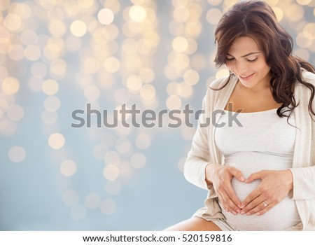 pregnancy, love, people and expectation concept - happy pregnant woman making heart gesture over holidays lights background