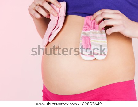 pregnancy, maternity and health concept - belly of a pregnant woman with two pairs of baby mittens