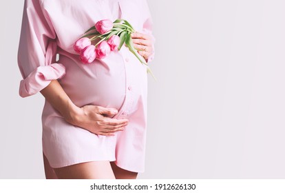 Pregnancy, Motherhood, Mother's Day Holiday Concept. Young Woman In Maternity Shirt Dress With Tulips Flowers Holds Hands On Belly. Beautiful Pregnant Woman Waiting For Baby Birth.