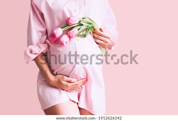 Pregnancy,\
Motherhood, Mother\'s Day Holiday concept. Young woman in maternity\
shirt dress with tulips flowers holds hands on belly. Beautiful\
pregnant woman waiting for baby\
birth.