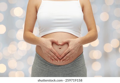 pregnancy, motherhood, people, love and expectation concept - close up of happy pregnant woman making heart shape hand sign on her bare tummy over holiday lights background