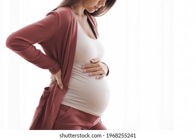 Pregnancy Pain. Cropped Shot Of Young Pregnant Lady Feeling Unwell At Home, Millennial Female Standing Against Window, Touching Belly And Lower Back, Suffering Prenatal Contractions, Free Space