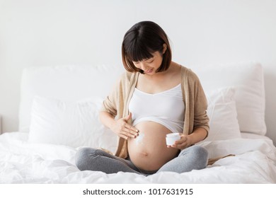 pregnancy, people and maternity concept - happy pregnant asian woman applying stretch mark cream to belly